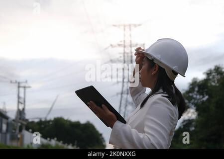 Asian female electrical engineer working on laptop near high voltage pole, inspecting power grid. Wind Turbine Engineer for Electric Power Generation. Stock Photo