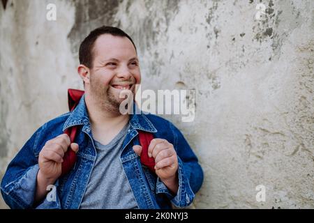 Portrait of happy young man with Down sydrome with backpack in street, smiling. Stock Photo