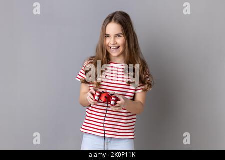 Portrait of little girl wearing striped T-shirt holding in hands red gamepad joystick, looking at camera with optimistic expression, enjoying game. Indoor studio shot isolated on gray background. Stock Photo