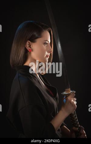 Portrait in profile of a young woman who holds a katana in her hands in front of her on a black background Stock Photo