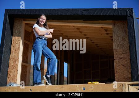 Happy woman on construction site standing on terrace at unfinished wooden frame house in the Scandinavian style barnhouse on sunny day, pointing at future building. Stock Photo