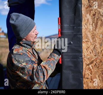 Male builders using red spirit level and measuring vertical surface of new wall, installing waterproof membrane. Men workers building wooden frame house. Carpentry and construction concept. Stock Photo