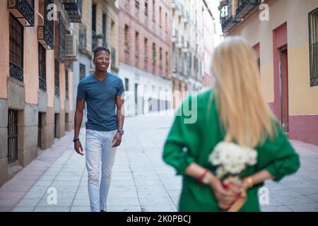 Selective focus on a african man walking towards a woman hiding a bouquet of flowers Stock Photo
