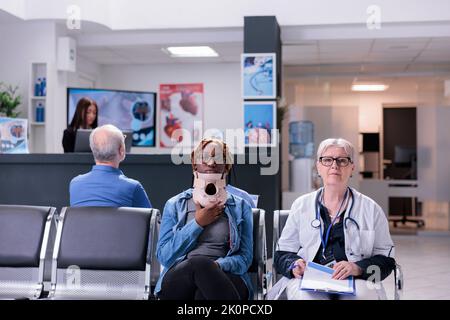 Portrait of person with neck collar brace sitting in waiting room at clinic doing consultation with senior doctor. Woman wearing cervical foam after accident injury talking to medic about healthcare. Stock Photo