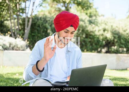 Smiling and friendly hindu male freelancer wearing national turban using laptop for video meeting sitting on the lawn outdoors. Indian student guy waving, studying remotely. Video call concept Stock Photo
