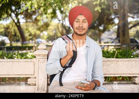 Portrait of handsome purposeful Indian male student with backpack holding phone and looking at camera in campus area, hindu freelancer man in traditional turban headwrap Stock Photo