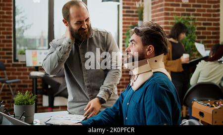 Business man in pain wearing cervical neck foam at office job, working on laptop and being hurt. Male employee with medical collar brace suffering after physical accident injury at work. Stock Photo