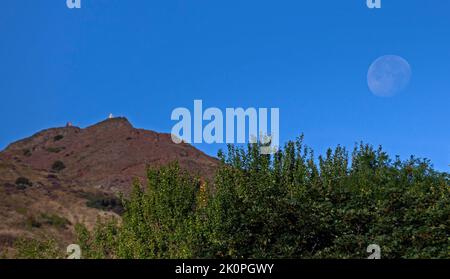 Holyrood Park Edinburgh, Scotland, UK 13th September 2022. Waning Gibbous Moon 90% appearing in the morning sky to the right of Arthur's Seat summit  with a backdrop of blue sky. Temperature 13 degrees centigrade. Credit/Arch White/alamy live news
