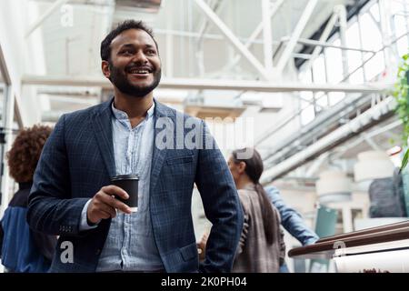 Smiling indian businessman drinking coffee and looking side while standing in office Stock Photo