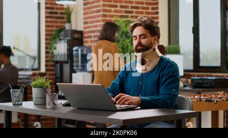 Hurt businessman wearing cervical neck brace at office job, working on project with pain and injury. Person with physical tension and collar foam trying to cure after accident, suffering at work. Stock Photo