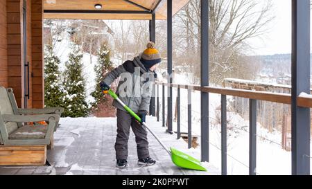 Involving children in housework concept. The boy cleans the snow on the terrace of his house with a big shovel.  Little helpers shovel snow. Family ac Stock Photo