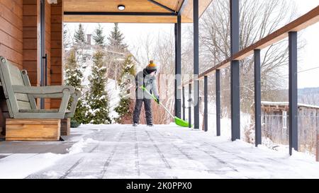 The boy cleans the snow on the terrace of his house with a big shovel. Family lifestyle in winter. The child is doing household chores. Kid shoveling Stock Photo
