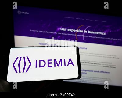 Person holding cellphone with logo of technology company IDEMIA France SAS on screen in front of business webpage. Focus on phone display. Stock Photo