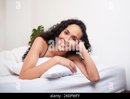 A beautiful young woman lies on her stomach on pillow in bed, smiling and happy Stock Photo