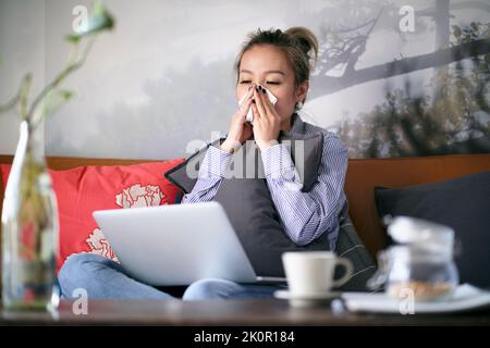 mature asian woman catching a cold, with running nose, working from home sitting on sofa using notebook computer Stock Photo