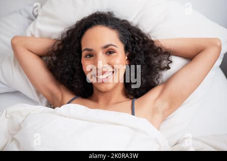 A young multi-ethnic woman lies with arms behind head in bed smiling relaxing Stock Photo