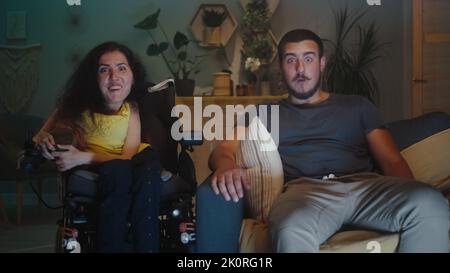 Shocked woman with physical disability in a wheelchair and man wondering and looking at each other, while watching TV show together at home in the evening Stock Photo