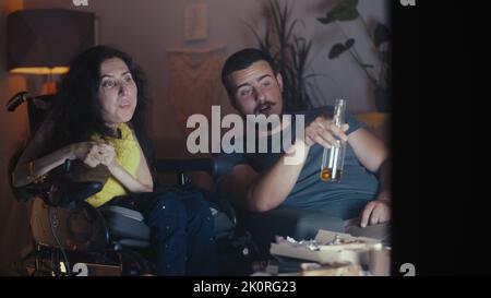 Woman with disability in a wheelchair talking and watching movie or sport games on TV in the evening at home, with her husband who drinking beer Stock Photo