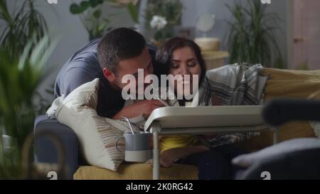 Husband covering his wife with disability with blanket while she sitting on sofa at the table and drawing using a digital pencil Stock Photo