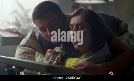 Woman with a disability sitting on a couch at the table, using digital tablet computer and pencil while her husband entering the room and covering her with a blanket Stock Photo