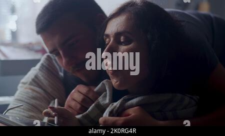 Woman with a disability talking to her husband sitting on a couch in a blanket and showing illustration app on a digital tablet computer Stock Photo