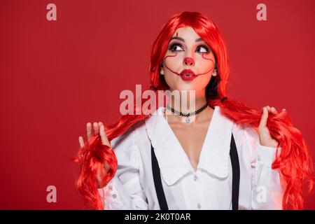 tricky woman with clown makeup holding bright ponytails and looking up isolated on red Stock Photo