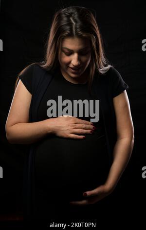 Young brunette woman holding her pregnant belly, studio light used. Stock Photo