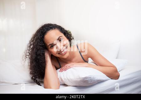 A beautiful young woman lies on her stomach on pillow in bed, smiling and happy Stock Photo