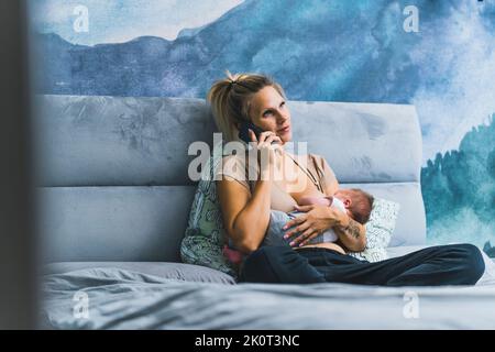 Caucasian beautiful young mother looking at the camera and holding her adorable newborn baby boy wrapped in a blanket, putting him to sleep, standing near the window at home. High quality photo Stock Photo