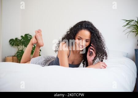 A young multi-ethnic woman lies on her stomach on bed, texting friends on phone Stock Photo