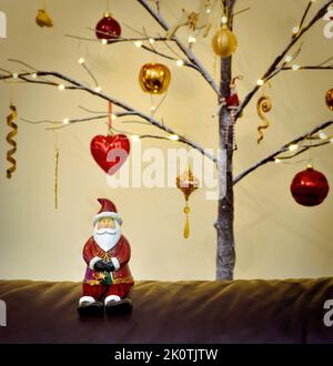 Father Christmas figure sat in front of a Twig Tree Christmas Tree Decorated in traditional red and gold Stock Photo