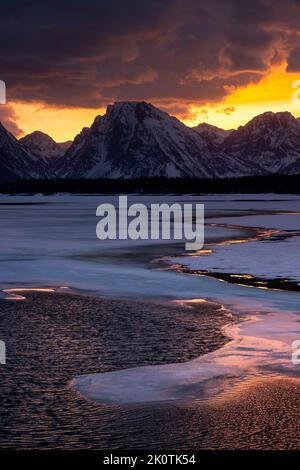 Thawing ice in the unusually low waters of Jackson Lake reflecting a colorful sunset behind Traverse Peak and the Teton Mountains. Grand Teton Nationa Stock Photo
