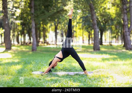 Young pregnant woman practicing yoga, doing a variation of Adho Mukha Svanasana, downward facing dog pose, exercising in the park on a sunny summer mo Stock Photo