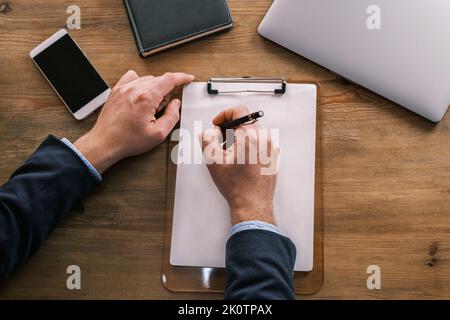 Top view of male hands writing something in a blank letterhead pinned to a clipboard. Businessman working at the table next to laptop, notepad and Stock Photo