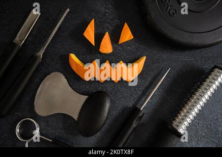 Gym dumbbell barbell weight plate with Halloween pumpkin pieces cut out. Carved face elements and carving tools, spoon gutter, saw blades. Stock Photo