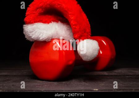Heavy red dumbbell and Santa Claus hat. Exercise equipment as a Christmas gift idea. Healthy fitness lifestyle holiday season concept composition. Stock Photo