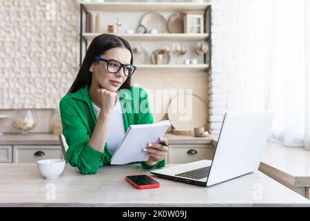 A young businesswoman in glasses sits at home in the kitchen with a laptop and conducts an online remote interview for the job of new employees. Listens carefully, holds a notebook, takes notes. Stock Photo