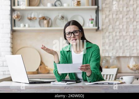 Divorce. A young beautiful woman received divorce papers. Shocked, upset. Sitting in the kitchen at home, wearing glasses and a green shirt, holding a letter in his hands. Stock Photo