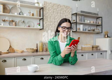 In love and happy young beautiful woman sends and receives love messages on the red phone from a loved one, from a boyfriend. Standing in the kitchen at home in glasses and a green shirt. Stock Photo