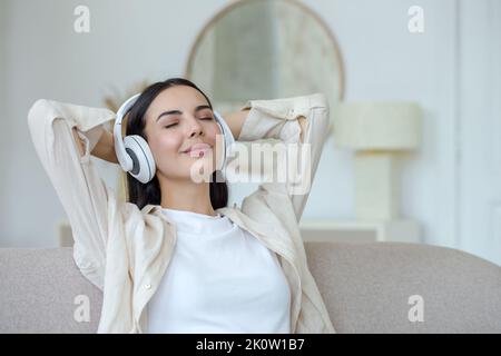 Young beautiful woman listening to music in white headphones, sitting on the sofa in the room, closed her eyes, put her hands behind her head, relaxed, resting. Stock Photo