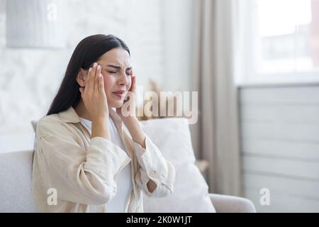 Headache. A young beautiful woman feels a strong pain in her head, holds her head with her hands, frowned in pain, closed her eyes. Sitting on the couch at home, needs help. Stock Photo