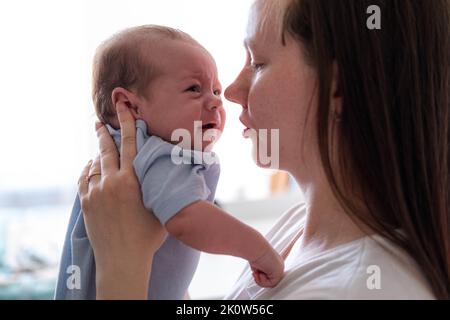 Mommy tries to calm the crying baby, shakes him in her arms, Dissatisfied, crying, upset, sad Newborn baby boy on mother hands at home. Stock Photo