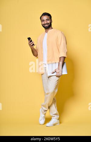 Indian man isolated on yellow holding laptop using mobile. Full length portrait Stock Photo
