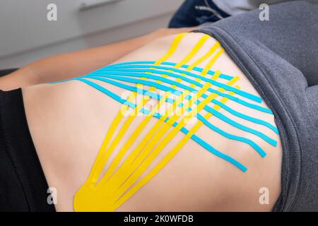 Flat lay above view applying blue adhesive elastic therapeutic tape on female abdominal muscle of young adult slim sporty woman lying on couch doctor Stock Photo