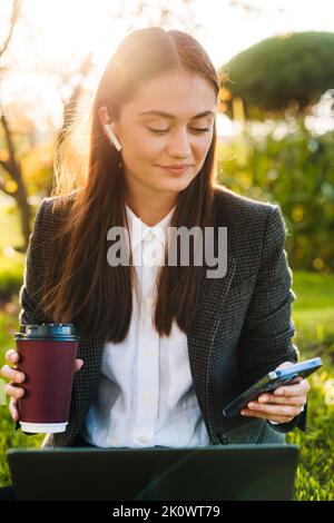 Portrait of a happy smiling working woman sending text messages from phone, outdoors, using laptop enjoying coffee on green grass. Internet Stock Photo