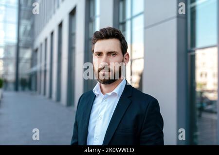 Headshot of serious attractive confident young caucasian guy with beard in suit looks at camera on street Stock Photo