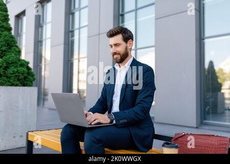 Cheerful attractive young caucasian guy manager with beard in suit typing on laptop, sits on bench Stock Photo