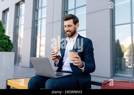 Successful business and lunch. Smiling handsome young caucasian man with beard in suit watches video on laptop Stock Photo