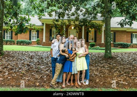 A large and blended and mixed happy family standing in front of their home in a group hug or embrace Stock Photo