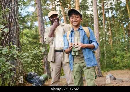 Two African American cheerful hikers with backpacks standing in pine tree forest and having fun while one of them holding binoculars Stock Photo